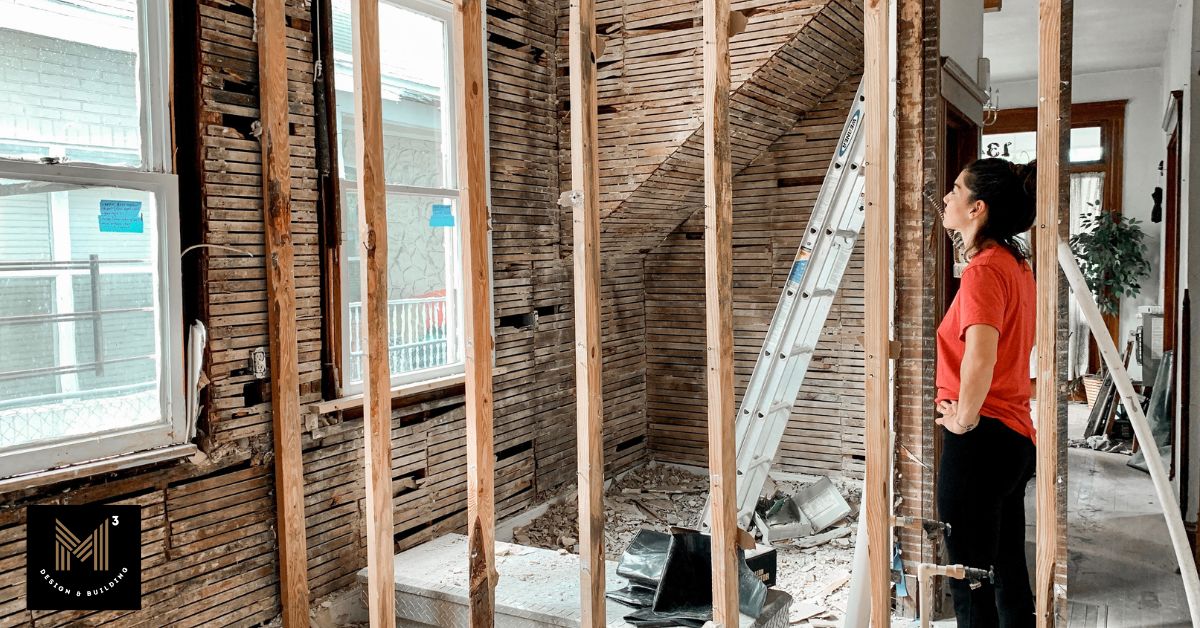 Partially demolished residential interior with exposed wall framing and a homeowner reviewing ongoing construction, illustrating value engineering decisions made during active construction when project changes require greater coordination.